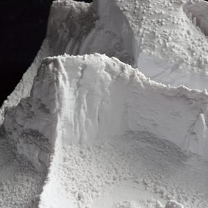 May include: A detailed, close-up view of a white, textured model of a mountain range. The model features rugged peaks, cliffs, and a granular surface, suggesting a realistic depiction of mountainous terrain. The lighting highlights the textures and contours.