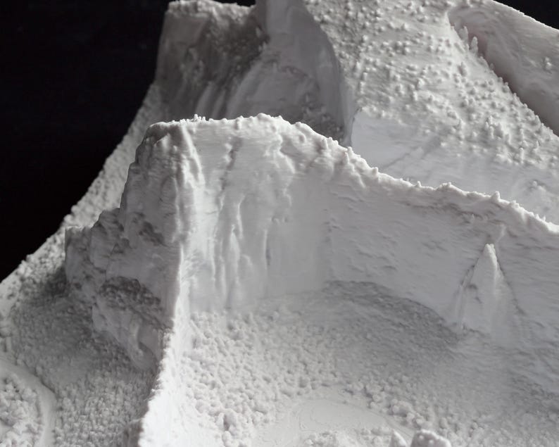May include: A detailed, close-up view of a white, textured model of a mountain range. The model features rugged peaks, cliffs, and a granular surface, suggesting a realistic depiction of mountainous terrain. The lighting highlights the textures and contours.