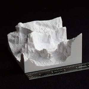May include: A white 3D-printed model of a canyon landscape, featuring detailed rock formations and a textured surface. A black ruler is placed next to the model for scale, showing measurements in inches.