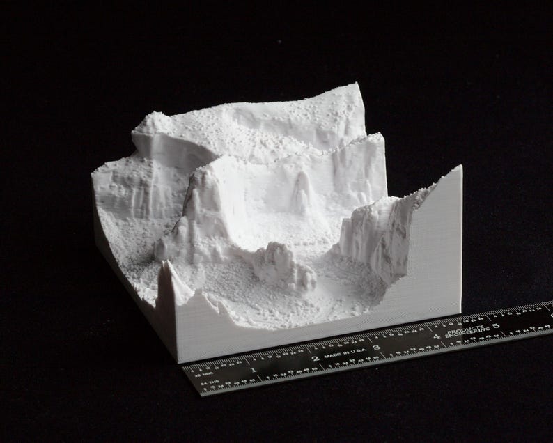 May include: A white 3D-printed model of a canyon landscape, featuring detailed rock formations and a textured surface. A black ruler is placed next to the model for scale, showing measurements in inches.