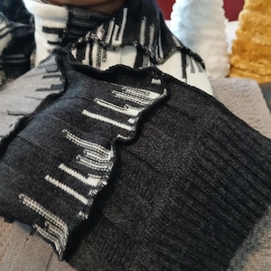 May include: Close-up of a black and gray knit scarf with a unique, geometric pattern. The scarf has a textured appearance and a contrasting white and black design along the edges. The scarf is part of a winter apparel collection.