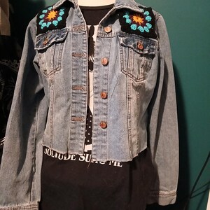 May include: Light blue denim jacket with crochet patches on the shoulders. The patches feature a turquoise and yellow floral design on a black background. The jacket has button closures and pockets. A black t-shirt with white text is visible underneath.