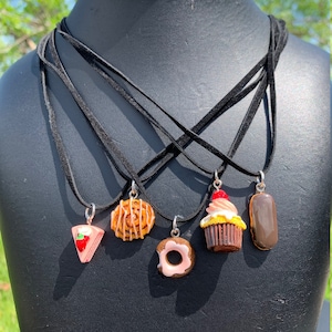 Könnte beinhalten: Ein Set aus fünf schwarzen Wildlederketten mit verschiedenen Essens-Anhängern. Die Anhänger umfassen ein Erdbeer-Shortcake-Stück, eine Zimtschnecke, einen Donut, einen Cupcake mit einer Kirsche oben drauf und einen mit Schokolade überzogenen Long John.