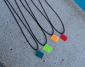 Puede incluir: Cinco collares con cordones negros y colgantes rectangulares de colores. Los colgantes son translúcidos y presentan un diseño pixelado. Los colores de los colgantes son verde azulado, verde, amarillo, naranja y rojo.