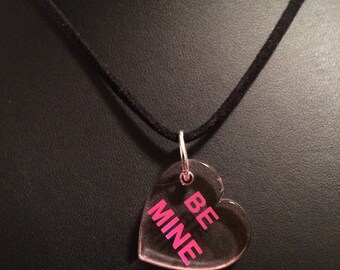 Op de afbeelding: Een zwarte suède ketting met een heldere hartvormige hanger waarop in roze letters "BE MINE" staat.