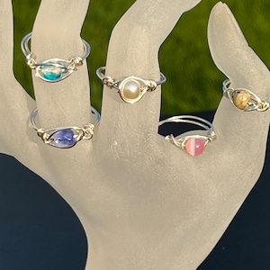May include: Five silver wire wrapped rings with different colored stones. The stones are blue, white, pink, brown, and green.