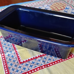 May include: A rectangular, navy blue ceramic baking dish with rounded edges. The dish is empty and sits on a yellow tablecloth with a red, white, and blue geometric pattern. The dish is approximately 12 inches long.