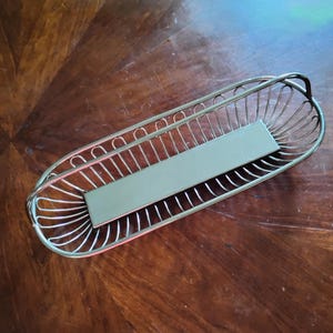 May include: An elongated, oval-shaped silver-toned metal basket featuring a wire-frame design with vertical slats and a solid, flat rectangular base. The basket includes a curved handle on each end, creating a structured, open-work container.