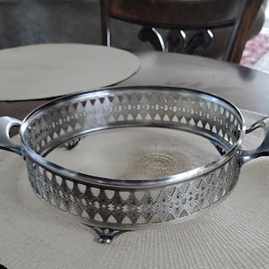 May include: Silver-colored serving dish with a decorative pierced design around the rim and two handles. The dish has a round shape and sits on three small feet. The interior is smooth and reflective, suitable for serving food or displaying items.