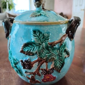 May include: A turquoise ceramic jar with a lid, featuring a blackberry and leaf design. The jar has two dark brown handles and a matching lid with a leaf-shaped knob. The design includes green leaves, brown branches, and black and red berries.