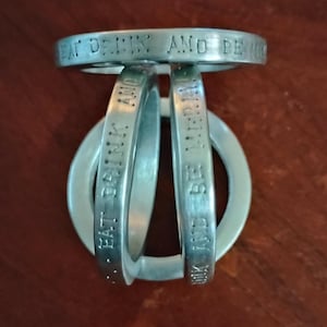May include: A silver-colored metal decorative wine bottle holder. The holder has a circular top and base, with three curved bands connecting them. The words "EAT DRINK AND BE MERRY" are engraved on the bands.