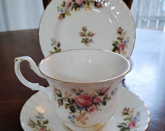 May include: A white porcelain teacup and saucer set with a matching side plate, featuring a delicate floral pattern of pink roses and small blue flowers with gold trim along the scalloped edges of the pieces and the teacup handle.
