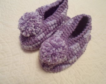 May include: A pair of handmade crochet slippers in shades of light and dark purple. Each slipper features a thick, textured yarn construction with a prominent, round pom-pom attached to the top of the toe area and a low-cut opening for the foot.