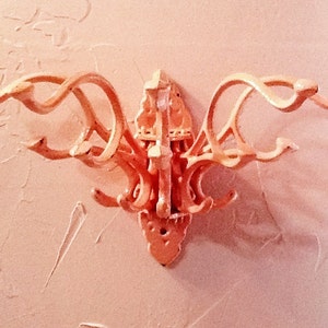 Puede incluir: Un perchero decorativo de pared con múltiples brazos y ganchos curvos. El perchero está hecho de un material de color claro, posiblemente madera o metal, y está montado en una pared texturizada. El diseño presenta detalles ornamentados y una disposición simétrica.