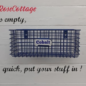 May include: A cobalt blue wire basket with a rectangular shape, mounted on a white wooden wall. The basket has a label that reads "Cobalt". Text on the wall says "it's empty, quick, put your stuff in!"