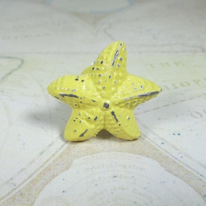 May include: A yellow starfish-shaped decorative object with a distressed finish. The five-pointed star has a textured surface and dark accents. This nautical-themed item could be used as a cabinet knob or decorative accent.