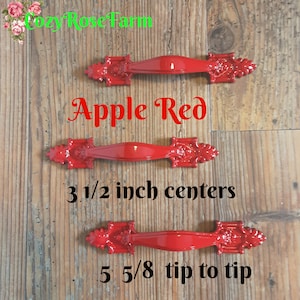 May include: Three apple red cabinet pulls with ornate end details. The text "Apple Red" is displayed above the pulls. The pulls are 3 1/2 inch centers and 5 5/8 tip to tip.