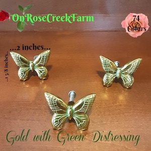 May include: Three gold-colored butterfly cabinet knobs with green distressing. Each butterfly measures approximately 2 inches wide and 1 5/8 inches high. The text "On Rose Creek Farm" and "74 Colors" are also visible.