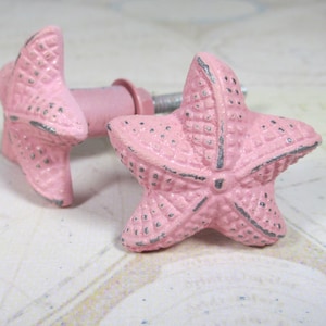 May include: Two pink starfish-shaped cabinet knobs with a distressed finish. The knobs have a metal screw and a pink stem. The starfish design has textured details and a slightly weathered appearance, adding a coastal or nautical touch to furniture.