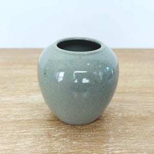 May include: A small, round, celadon-colored ceramic vase. The vase has a wide opening and a smooth, glossy finish. It sits on a light brown wooden surface. The vase is a decorative item.