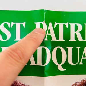 May include: A section of a green poster with white text that reads "ST PATRICK HEADQUAR". A finger points to the poster. The poster is creased at the centre.