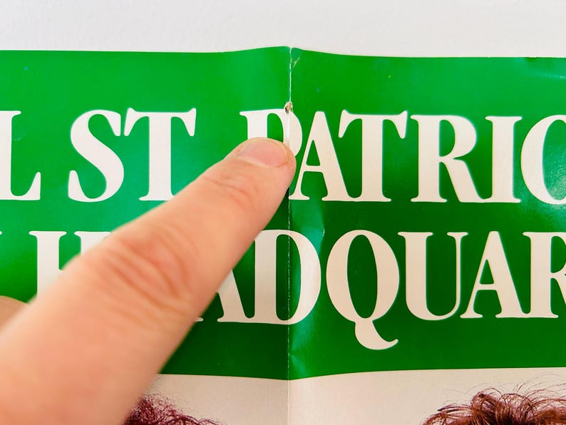 May include: A section of a green poster with white text that reads "ST PATRICK HEADQUAR". A finger points to the poster. The poster is creased at the centre.