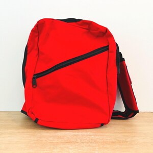 May include: A bright red messenger bag with a black zipper and trim. The bag has a single shoulder strap and a zippered front pocket. The bag is sitting on a wooden surface.