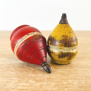 May include: Two antique wooden spinning tops. One is red with a white stripe, the other is yellow with brown accents. Both have a metal tip. The tops are resting on a light brown wooden surface.