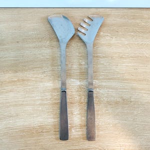 May include: Two stainless steel salad servers. One server has a flat, wide head, and the other has a slotted head with four tines. Both servers have long, rectangular handles with a darker finish. The servers are on a light wood surface.