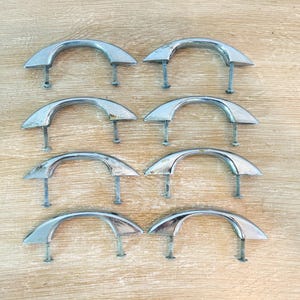 May include: A set of eight silver-toned metal drawer pulls arranged in two columns. Each handle features a smooth, arched design with two vertical mounting screws extending from the underside, showcasing a polished, reflective metallic finish.