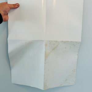 May include: A folded white paper with creases and discolouration. The paper is held by a hand, revealing its folded structure. A piece of tape secures the top. The paper's surface shows signs of age and wear.