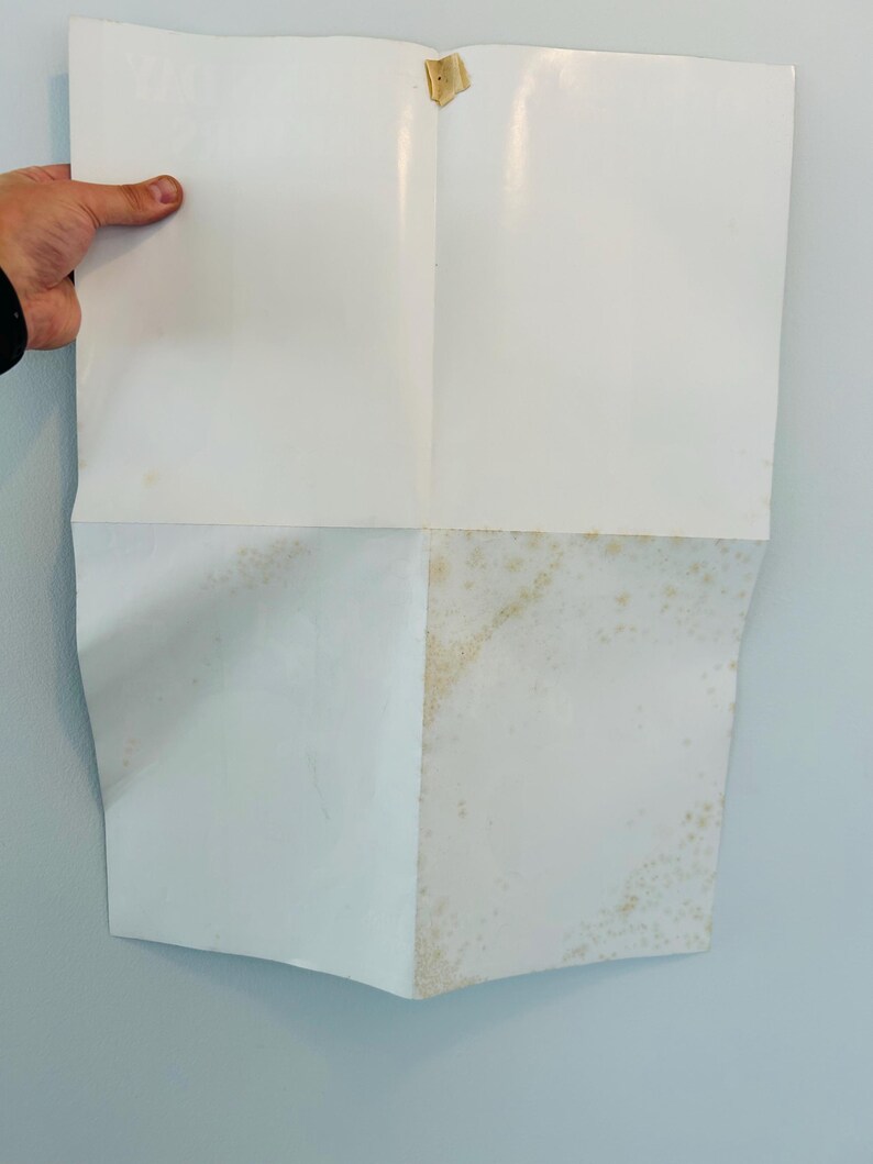May include: A folded white paper with creases and discolouration. The paper is held by a hand, revealing its folded structure. A piece of tape secures the top. The paper's surface shows signs of age and wear.