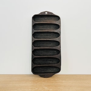 May include: A vintage cast iron cornbread pan with six individual molds. The pan is dark brown and has a rustic look.