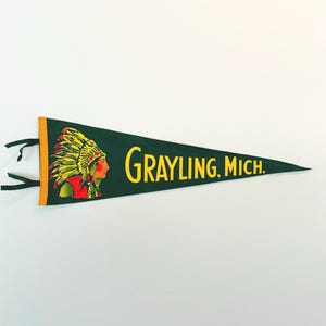 May include: A dark green pennant with the text "GRAYLING, MICH." in yellow. The pennant features an illustration of a Native American chief in a headdress. The pennant has a yellow border and black ties.