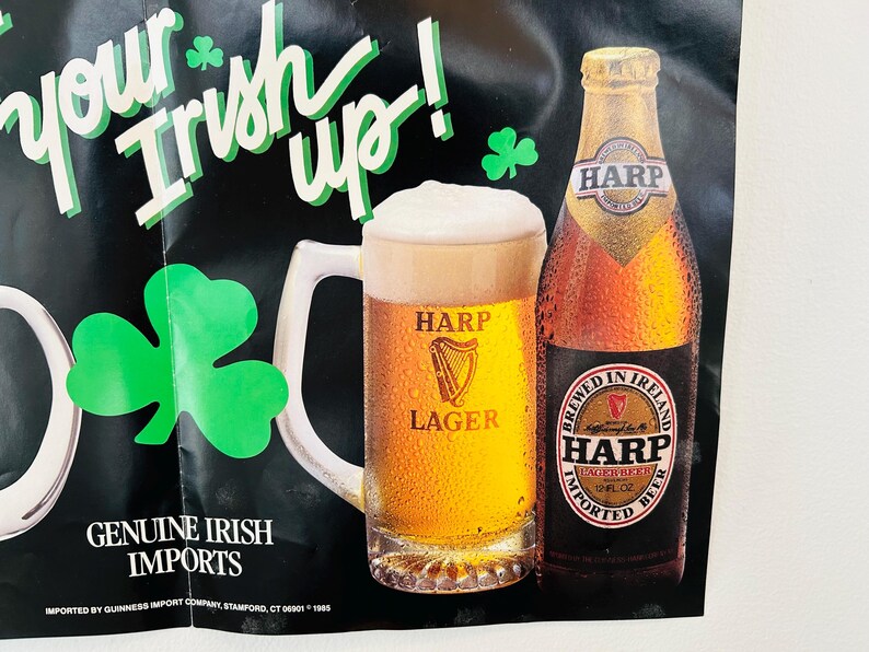 May include: Poster advertising Harp Lager beer, featuring a pint of beer and a bottle. The text reads "Your Irish up!" with shamrock decorations. The poster also states "Genuine Irish Imports."