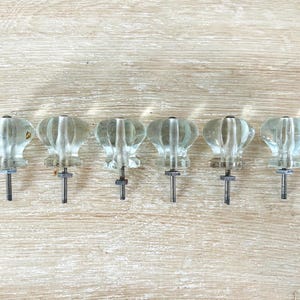 May include: Six clear glass cabinet knobs with a ribbed, flower-like design. Each knob has a metal screw post for mounting. The knobs are arranged in a row on a light wood surface.