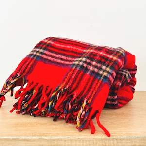 May include: A folded red plaid blanket with a fringed edge. The blanket features a bold red base color with a classic plaid pattern in black, white, and blue. The blanket is resting on a light brown wooden surface.