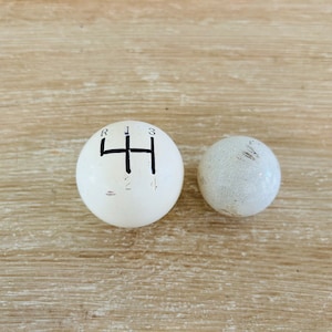 May include: Two spherical gear shift knobs. One is white with a black shift pattern diagram, including R, 1, 2, 3, and 4. The other is a smaller, textured, off-white sphere.