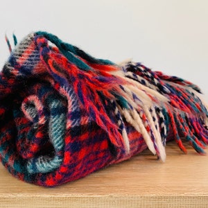 May include: Rolled plaid blanket in shades of red, teal, navy, and cream. The blanket has a fringed edge and is displayed on a wooden surface. The plaid pattern is visible throughout the blanket.