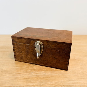 May include: A dark brown wooden box with a silver latch and a handle. The box is closed and sitting on a light brown wooden surface.