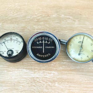 May include: Three circular analog gauges. The left is a black Weston Volts D.C. meter with a white face. The center is a chrome-rimmed ammeter with a black face labeled Discharge and Charging. The right is a silver Ames dial indicator gauge.