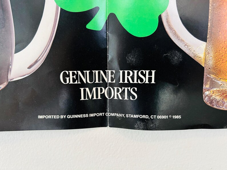 May include: A vintage advertisement featuring the text "GENUINE IRISH IMPORTS" in white on a black backdrop. A green shamrock and a pint of beer are visible. The advertisement is from 1985.