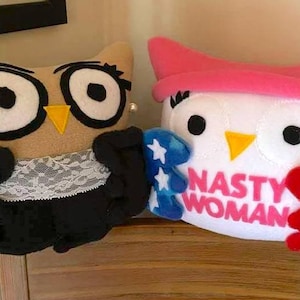 May include: Two plush owl dolls. One is tan with glasses, a pearl earring, and a black robe with a lace collar. The other is white with a pink hat and the words "NASTY WOMAN" on its chest. One holds a blue star-covered item, the other a red and white striped item.