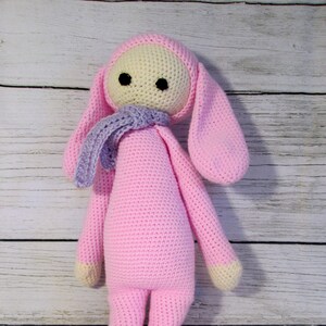 May include: A pink crocheted bunny doll with long ears and a purple scarf. The doll has a cream-colored face with black eyes.
