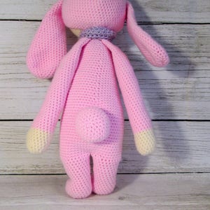 May include: A pink crocheted bunny rabbit toy with long ears and a small tail. The bunny is standing on a white wooden surface.