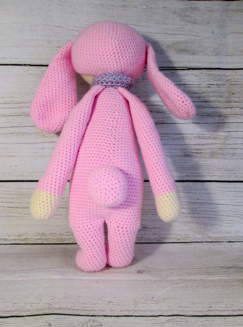 May include: A pink crocheted bunny rabbit toy with long ears and a small tail. The bunny is standing on a white wooden surface.