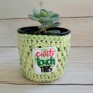 May include: A green crocheted plant pot with a white patch that says "Can't touch this" in red and black lettering. A succulent plant is growing in the pot.