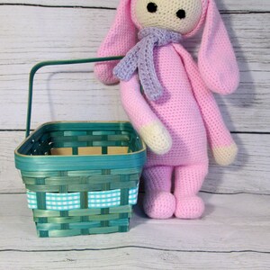 May include: A pink crocheted bunny doll with long ears and a purple scarf stands next to a teal woven basket with a white and teal checkered ribbon.