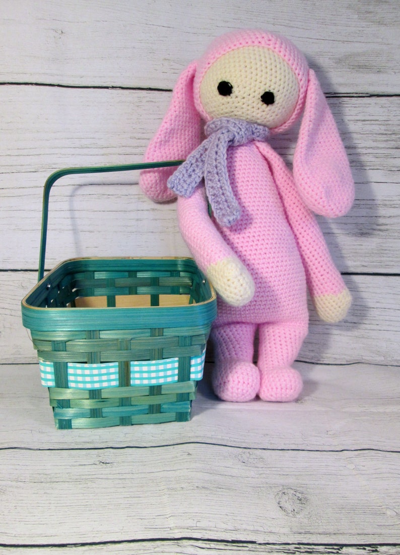 May include: A pink crocheted bunny doll with long ears and a purple scarf stands next to a teal woven basket with a white and teal checkered ribbon.