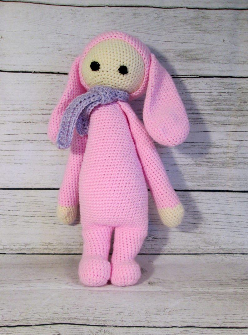 May include: A pink crocheted bunny doll with long ears and a purple scarf. The doll has a cream-colored face with black eyes.
